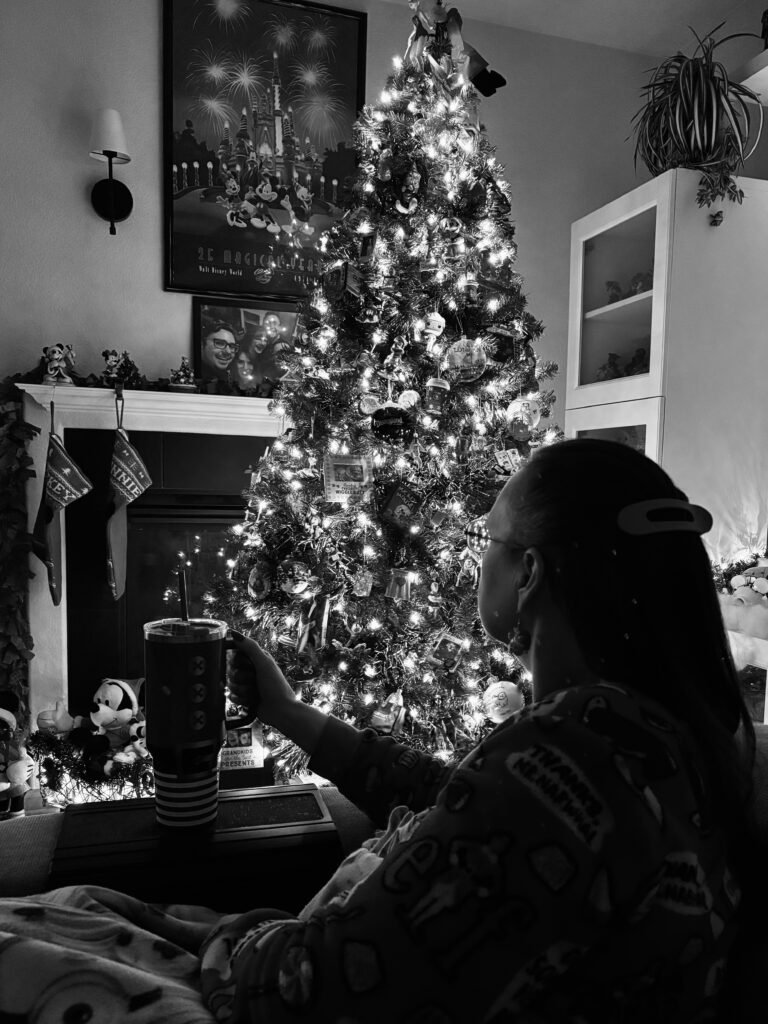 Black and white photo of a woman sitting quietly in her living room, holding a cup and looking at a decorated chirstmas tree glowing with lights, captured in a calm, reflective moment.