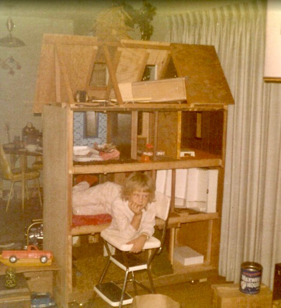 A childhood Christmas photo of Cathy standing next to the oversized Barbie dollhouse her father built by hand, a partially finished wooden house filled with imagination and love.