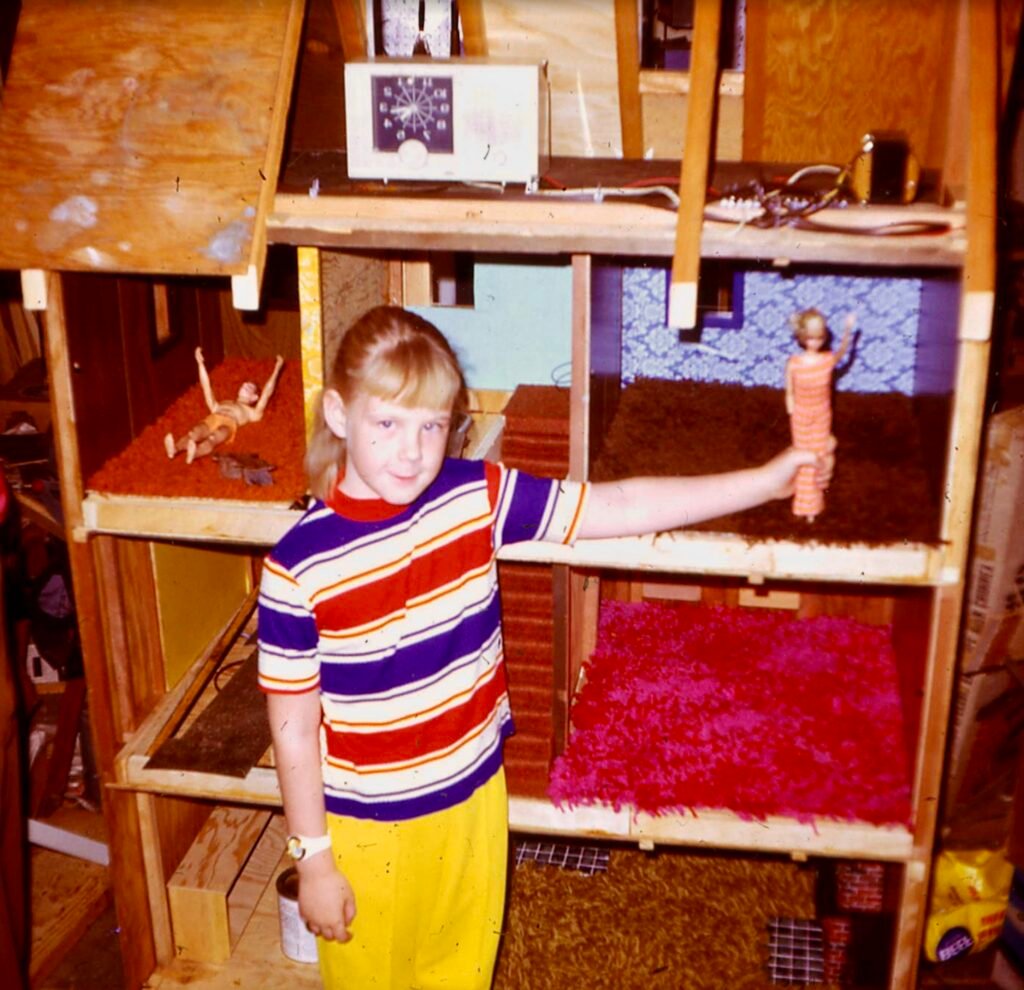 A childhood Christmas photo of Cathy standing next to the oversized Barbie dollhouse her father built by hand, a partially finished wooden house filled with imagination and lo