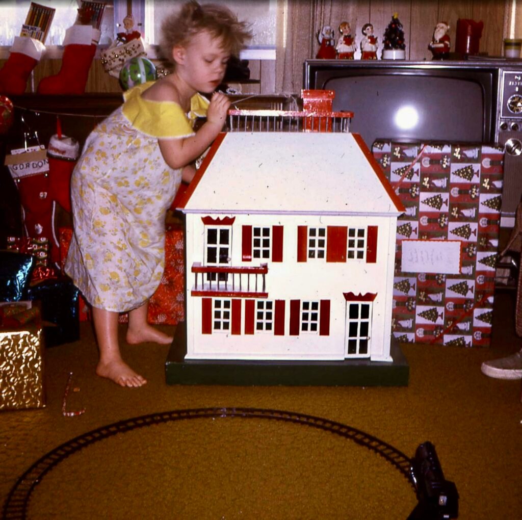 Little girl standing next to a white dollhouse with red trim that her grandfather made. 